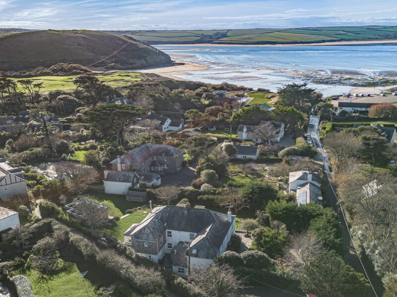 Panoramic aerial view of Cornish seaside village near the coast, featuring lush greenery, charming houses, and sandy beaches.