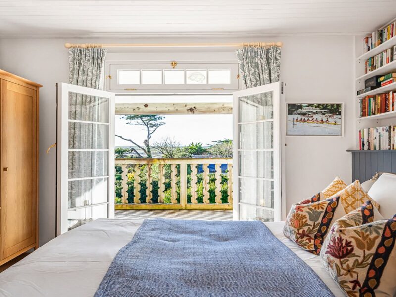 Coastal bedroom with sea view, wooden furniture, colourful cushions, and open French doors to a balcony.