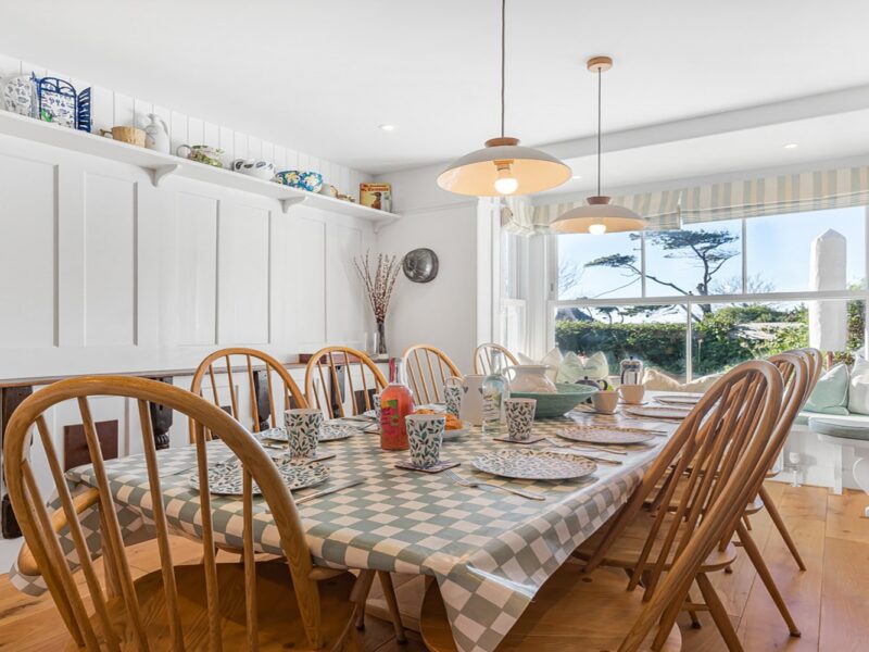 Bright, cosy coastal dining room with wooden furniture, white walls, and large windows overlooking gardens.