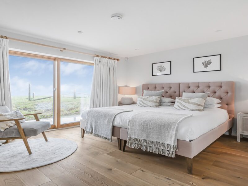 Coastal bedroom with pink upholstered bed, wooden flooring and sea view through large glass doors.