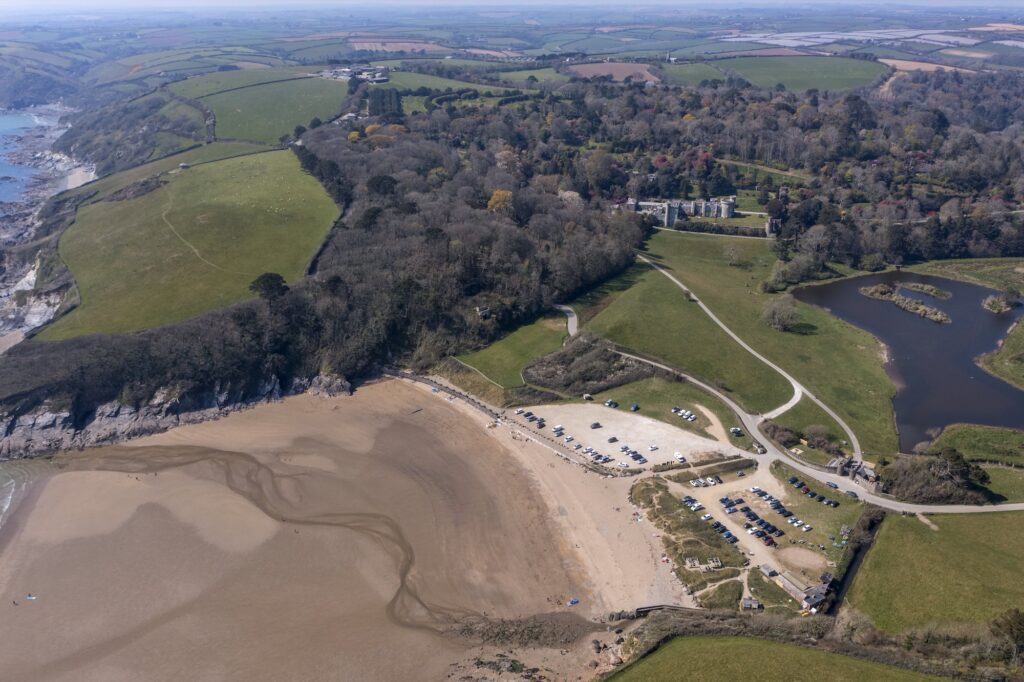 Aerial view of Cornish Secrets beach with parking lot, sandy shoreline, and surrounding green landsc.