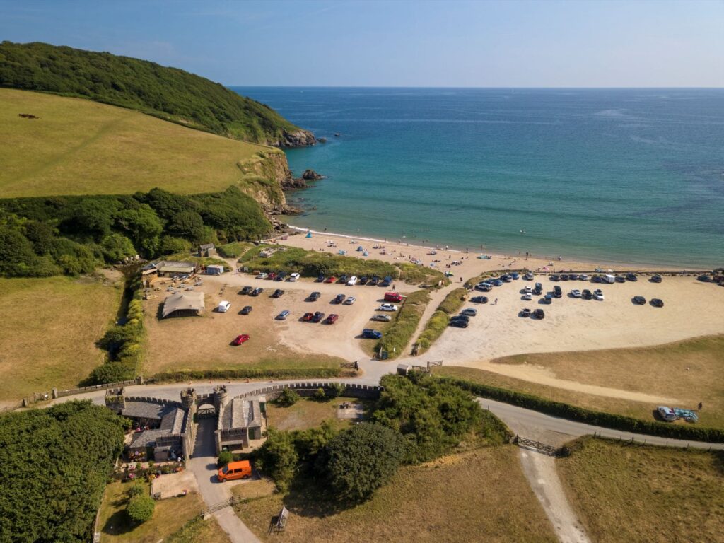 Aerial view of a sandy beach with turquoise waters, surrounded by green hills and a parking lot at C.