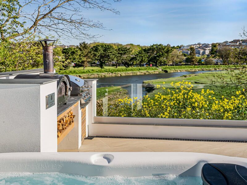 Hot tub overlooking a river with lush greenery and yellow wildflowers in Cornwall.